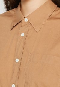 Camicia button-up marrone chiaro realizzata in tessuto liscio. Presenta un colletto a punta, bottoni bianchi e una tasca sul petto sul lato sinistro.
