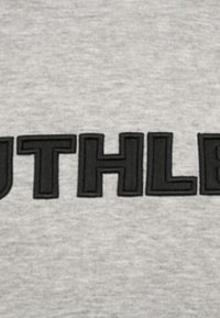 Grijs stof met grote, vette zwarte geborduurde letters die "RUTHLESS" spellen. De textuur lijkt zacht en casual.