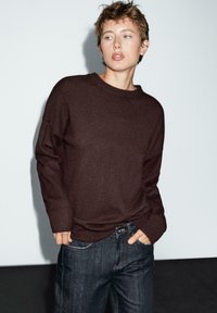Brauner Strickpullover mit rundem Ausschnitt und gerippten Bündchen, kombiniert mit dunklen Jeans. Einfaches Design; weiche Textur, keine Muster oder auffälligen Hardware.
