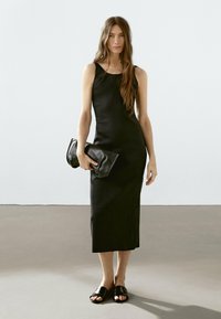 Abito nero senza maniche con una forma aderente, realizzato in tessuto liscio. Abbinato a una pochette nera e sandali piatti con apertura davanti. Design semplice.