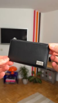 Rokas tur melnu Diesel ādas karšu turētāju ar rāvējslēdzēju dzīvojamā istabā ar augiem un televizoru fonā.