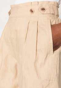 Beżowe lniane szorty z plisowanym wzorem, dwoma guzikami w pasie i bocznymi kieszeniami. Miękka faktura o naturalnym wykończeniu.