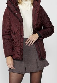 Veste matelassée bordeaux avec un design matelassé, des poches zippées et une capuche, associée à un col roulé crème et une jupe à carreaux.
