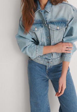 Kvinde iført lyseblå denimjakke med knappede brystlommer og medium blå højtaljede jeans, stående med armene over kors.