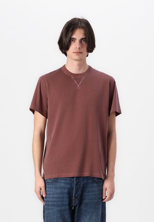 Camiseta de algodón marrón con mangas cortas, cuello redondo y aberturas laterales. Presenta un sutil logotipo en el pecho y una textura suave.