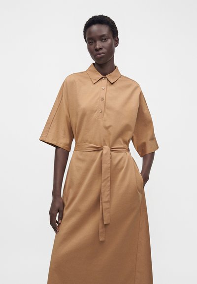 Femme portant une robe beige à manches courtes, boutonnée, avec un col et une ceinture nouée à la taille, debout devant un fond clair uni.