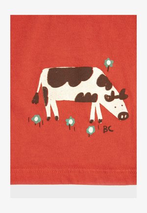 Vache dessinée en noir et blanc broutant parmi de petites fleurs bleues sur un fond de tissu rouge avec un ourlet cousu en bas.