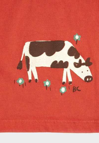 Vache dessinée en noir et blanc broutant parmi de petites fleurs bleues sur un fond de tissu rouge avec un ourlet cousu en bas.