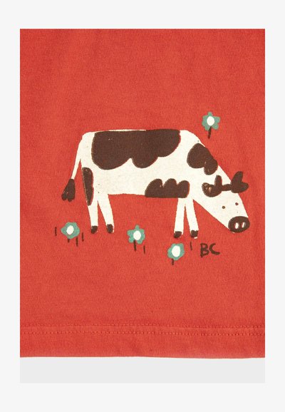 Vache dessinée en noir et blanc broutant parmi de petites fleurs bleues sur un fond de tissu rouge avec un ourlet cousu en bas.