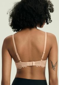Frau mit lockigem Haar, von hinten gezeigt, trägt einen beigen Spitzen-BH und schwarze Hosen, mit einem Feen-Tattoo auf ihrem rechten Oberarm.