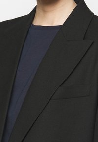 Svart blazer med spetsiga slag, skräddarsydd passform och bröstficka. Bärs över en marinblå skjorta som visar en slät textur och lager.