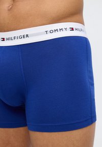 Κοντινό πλάνο ενός ατόμου που φορά μπλε μπόξερ Tommy Hilfiger με λευκή μέση και λογότυπο.