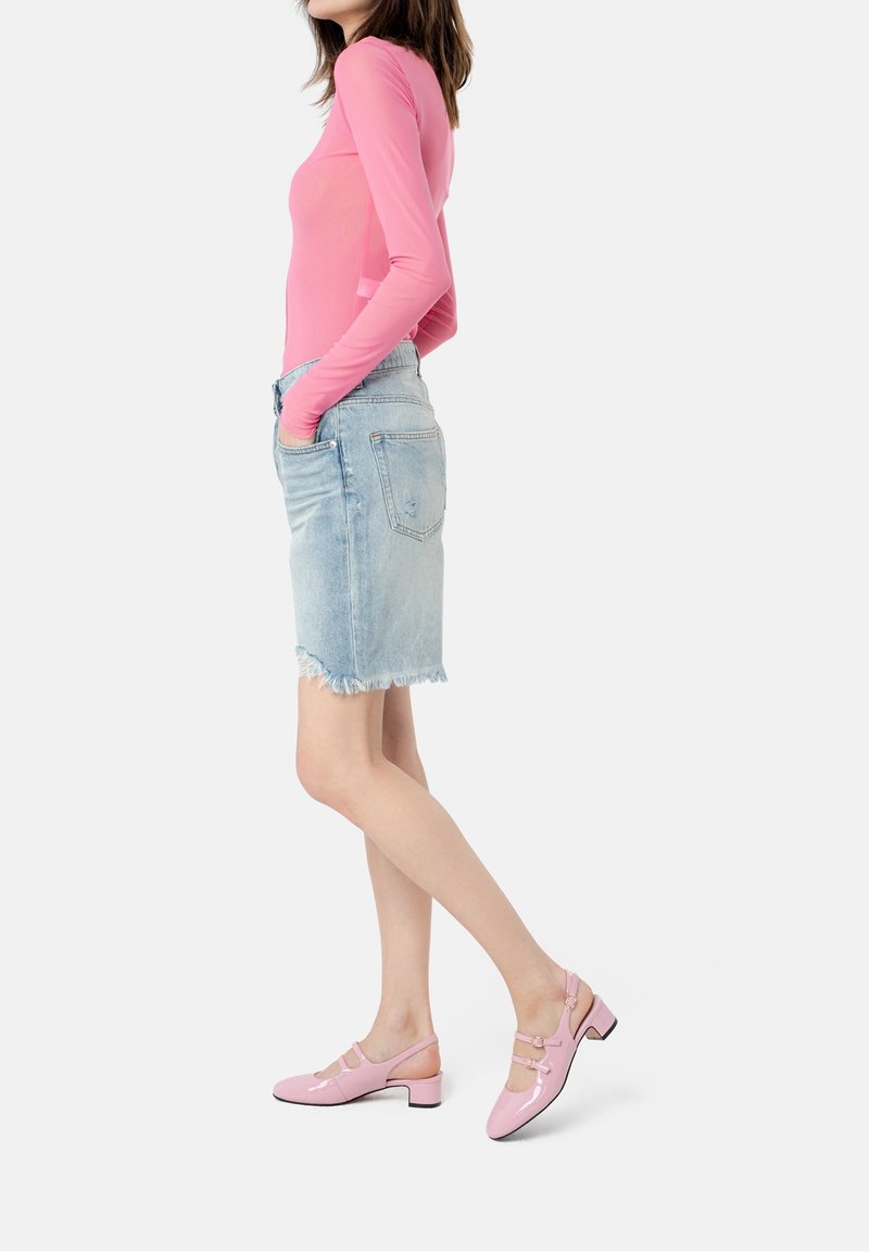 Femme portant un haut rose à manches longues, une jupe en jean bleu clair effilochée et des chaussures Mary Jane roses à talons bas, debout de profil.