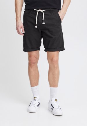 Schwarze Shorts mit umgeschlagenen Säumen, aus leichtem Stoff gefertigt. Verfügt über einen Kordelzugbund mit einem Knopfdetail. Getragen mit weißen Socken und Schuhen.