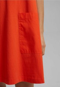 Röd bomullsklänning med en stor plånbok på vänster sida med synliga stygn och en fåll som når midjan.