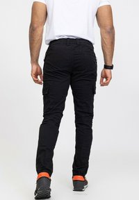 Pantalons cargo noirs avec une coupe slim, présentant des poches latérales et un tissu texturé. Portés avec des chaussures noires et des accents orange sur les semelles.