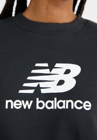 Felpa grigia scura con un grande logo bianco "New Balance" e un design stilizzato della lettera 'N'. Materiale in cotone, scollatura a girocollo.