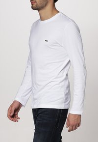 Balta ilga rankovė t-shirt, pagaminta iš medvilnės, su žaliu siuvinėtų krokodilo logotipu kairiajame krūtinės šone. Kasdienis fasonas su apvaliu iškirpimu.