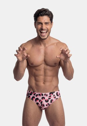 Homme souriant torse nu aux cheveux courts et foncés, portant un slip de bain rose et noir à motif léopard, posant avec les mains levées et les doigts recourbés.