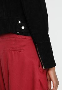 Svart mockajacka med silverdetaljer, sidogömfickor och en skräddarsydd passform, i kombination med röda tygbyxor med framfickor.