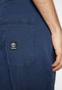 Pantaloni di cotone blu navy con tasca visibile e patch del logo Timberland. Presentano cuciture rinforzate e una texture liscia.