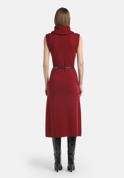 Ženska videna od zadaj nosi brezrokavnico v burgundski barvi midi obleko z zvitim ovratnikom in črne čizme do kolen s peto.