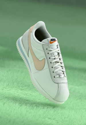 Biały sneaker Nike z beżowym znakiem Swoosh, białymi sznurowadłami i pomarańczowym logo Nike na języku, ustawiony na miękkiej zielonej powierzchni.