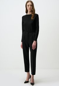 Schwarzes, geripptes Langarmoberteil, kombiniert mit maßgeschneiderten schwarzen Hosen mit Falten. Das Model trägt schwarze, spitze High Heels vor einem hellen Hintergrund.