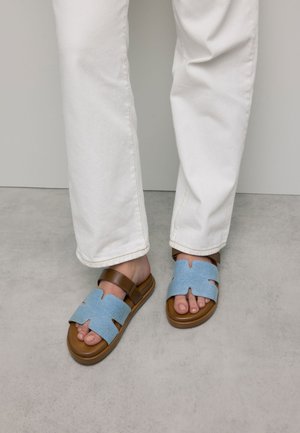 Blauwe stoffen sandalen met een open teenontwerp, een bruine verstelbare band en een stevige bruine zool, gedragen met witte broek.