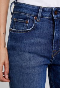 Närbild av blå denimjeans i midjan och övre låret med synliga framfickor och knapp, bredvid en hand med en pil-tatuering på underarmen.