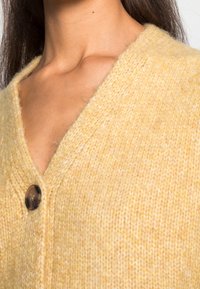 Żółty, dziergany kardigan z dekoltem w serek, pojedynczym ciemnym guzikiem i miękką teksturą. Zbliżenie na tkaninę i szczegóły dekoltu.