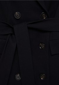 Manteau noir croisé avec six boutons foncés et une large ceinture en tissu nouée autour de la taille.