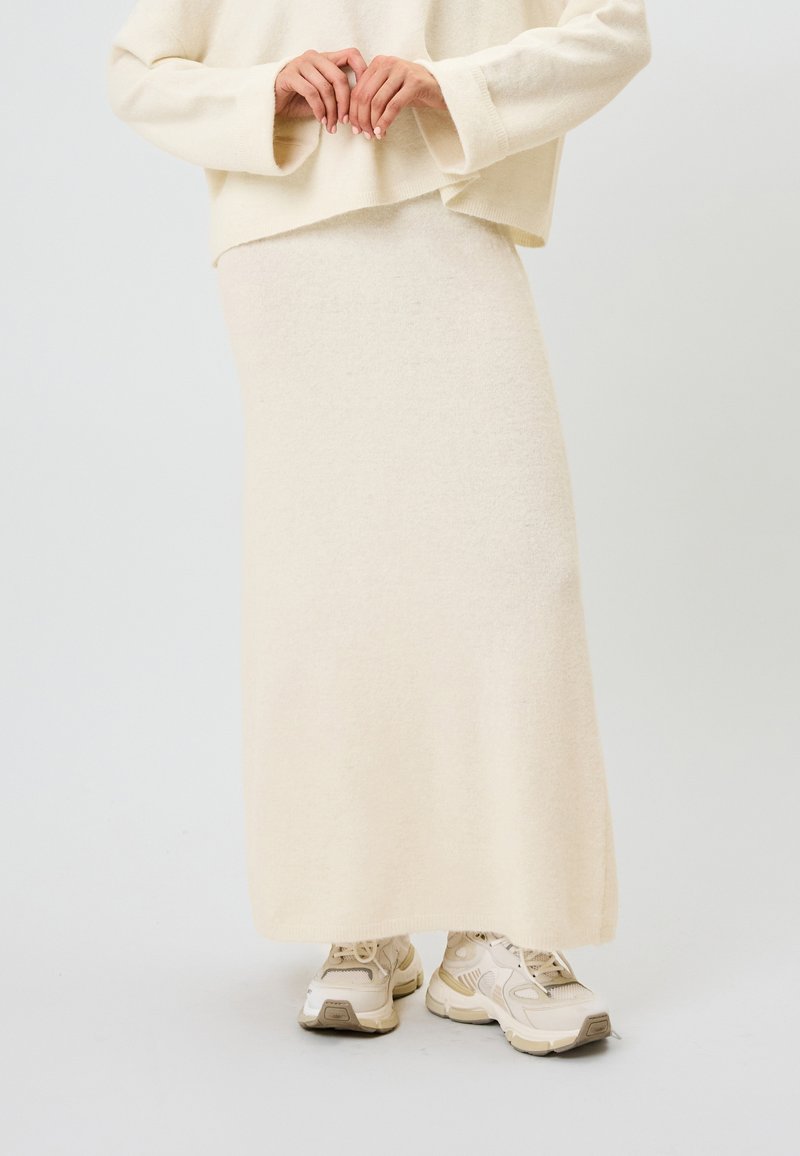 Beige neulottu maksihame, valmistettu pehmeästä materiaalista, jossa on suora siluetti ja hienovarainen tekstuuri, yhdistetty vaaleanvärisiin paksupohjaisiin lenkkareihin.