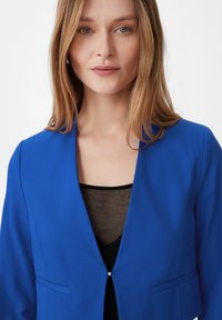 Niebieski żakiet z klapkowym kołnierzykiem, z pojedynczym zapięciem na haczyk, dwoma przednimi kieszeniami i gładką teksturą, nałożony na czarną bluzkę.