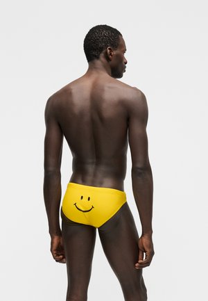 Άνδρας που στέκεται με την πλάτη στραμμένη, φοράει φωτεινά κίτρινα μαγιό με ένα μαύρο χαμογελαστό πρόσωπο τυπωμένο στο πίσω μέρος.