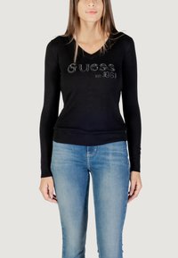 Maglione nero a maniche lunghe con scollo a V e logo testurizzato "Guess est. 1981". Design aderente, polsi e orlo a coste, abbinato a jeans blu chiaro.