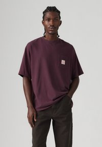 Donkerpaarse oversized t-shirt van katoen, met een borstzakje met een label. Gepaard met donkerbruine broek. Soepele textuur.