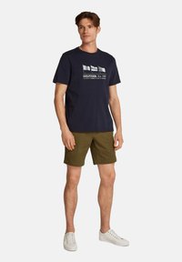 Camiseta de algodón azul marino con gráfico de bandera, combinada con pantalones cortos verde oliva. Modelo con zapatillas blancas, de pie contra un fondo sencillo.
