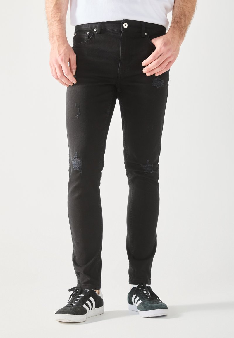 Fekete farmernadrág, slim-fit kivitelben, térden kopott részletekkel és öt zsebből álló stílussal. Fekete sportcipővel párosítva.