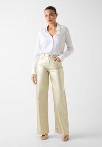 Wit overhemd met lange mouwen en kraag, gecombineerd met hooggetailleerde, wijde broekspijpen in metallic goud. Getextureerde stof met een subtiele glans.