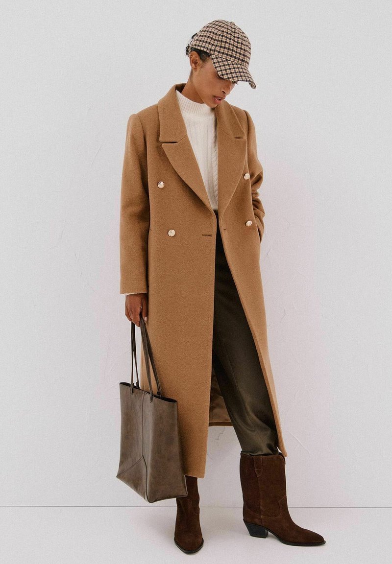 Lungo cappotto beige con bottoni doppi, maglione crema, pantaloni olivastro, stivaletti in camoscio marrone, e berretto a scacchi. Borsa a tracolla grigia.