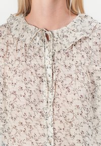 Blouse légère avec un motif floral, col à volants, fermeture à nouer à l'avant et design boutonnière en beige doux et couleurs atténuées.