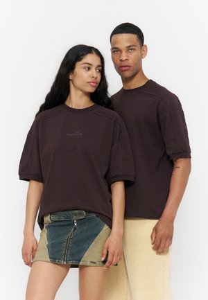 Junger Mann und Frau tragen passende dunkelbraune, übergroße T-Shirts; die Frau kombiniert sie mit einem zweifarbigen Jeansrock, der Mann mit hellen Hosen.