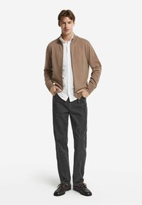 Hellbrauner Reißverschluss-Pullover aus weichem Material, getragen über einem weißen Hemd mit Kragen, kombiniert mit dunklen Jeans und schwarzen Schuhen mit braunen Schnürsenkeln.