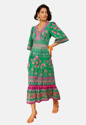 Kvinde iført en grøn blomster midi-kjole med pink og orange mønstre, orange sandaler, store guldsmykker og et orange armbånd.