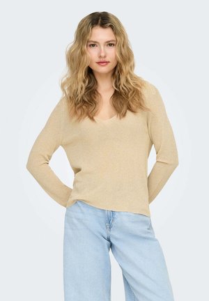 Donna con capelli biondi lunghi e ondulati, che indossa una maglia beige a maniche lunghe e jeans azzurri, in piedi con le mani dietro la schiena.