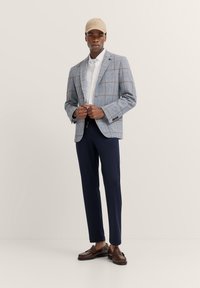 Hellblau karierter Blazer über einem weißen Hemd, kombiniert mit navyblauen Hosen und braunen Leder-Loafers. Das Modell trägt eine beigen Mütze.