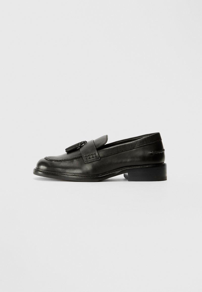 Mocassins en cuir noir avec un bout arrondi, dotés d'une sangle à l'avant et de coutures décoratives, reposant sur un petit talon carré. Texture lisse.