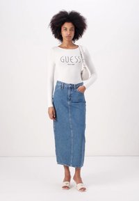 Valkoinen pitkähihainen paita, jossa on "GUESS"-logo, yhdistettynä korkeavyötäröiseen farkkuhameeseen. Asusteena on vaaleat liukuvaattet.