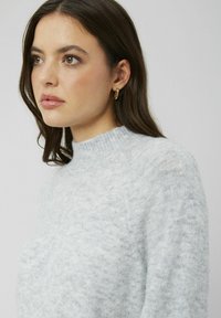 Pull en tricot texturé gris clair avec un col haut et des détails côtelés. Le modèle porte des boucles d'oreilles en or rondes.